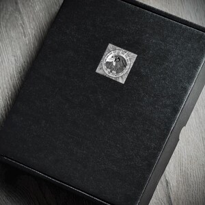 K&ouml;nnte beinhalten: Eine schwarze Box mit einem kreisf&ouml;rmigen Aufkleber auf der Oberseite. Der Aufkleber hat ein detailliertes Graustufen-Design mit einem Vogel und anderen Symbolen. Die Box steht auf einer Holzoberfl&auml;che.
