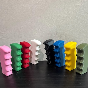 May include: A collection of eight 3D-printed, textured handles in various colors, including pink, green, red, white, black, blue, yellow, and olive green. The handles are arranged in a row, showcasing their unique design and color variations.