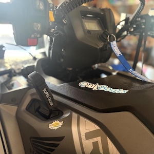 May include: Close-up of an ATV dashboard with a black and gray color scheme. Features include a digital display, a key with a lanyard, and the text "SUPER CRACKER" on a lever. The ATV also has the "OnlyBoats" logo.