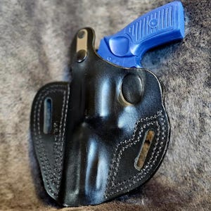 May include: A black leather holster with a blue handgun inside. The holster has stitching around the edges and a belt loop.