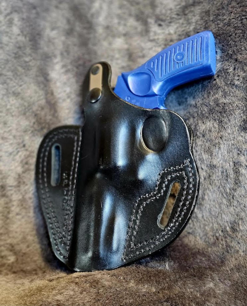 May include: A black leather holster with a blue handgun inside. The holster has stitching around the edges and a belt loop.