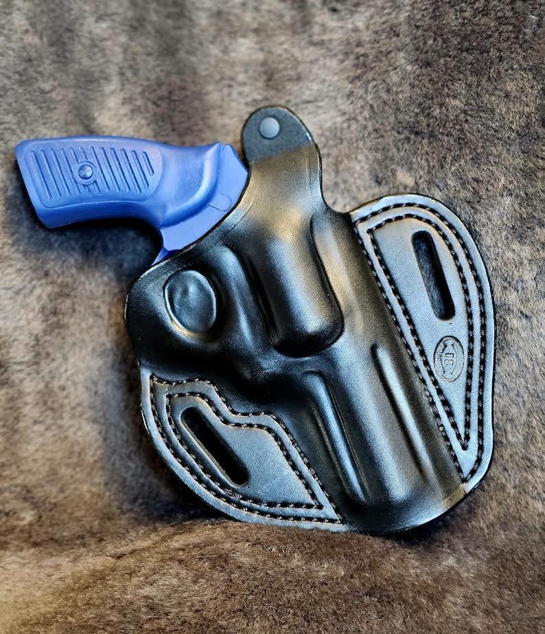 May include: A black leather holster with a blue handgun inside. The holster has a stitched design and a belt loop.