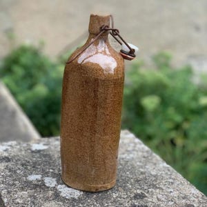 May include: A brown ceramic bottle with a speckled, textured finish. The bottle features a narrow neck and a metal swing-top closure mechanism with a white stopper and a rubber gasket, designed for sealing the opening at the top of the vessel.