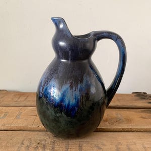May include: A ceramic pitcher with a rounded body and a curved handle, featuring a glossy glaze that transitions from a deep, dark navy blue at the top rim and neck to a mottled, earthy forest green hue across the lower portion of the vessel.