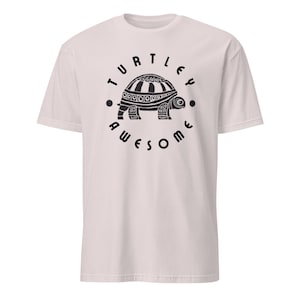 May include: Light pink t-shirt with a black graphic of a turtle inside a circle. The text "TURTLEY AWESOME" surrounds the turtle. The turtle design is detailed with patterns on its shell and body.