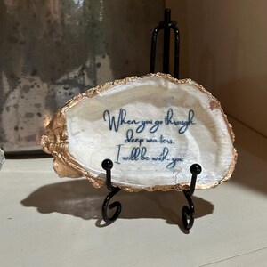 May include: A white oyster shell with gold trim on a black metal stand. The shell has a blue inscription that reads "When you go through deep waters, I will be with you."