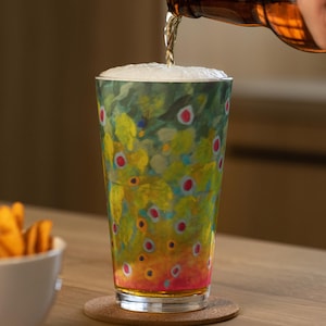 May include: A pint glass with a colorful, abstract design is being filled with beer from a brown bottle. The glass sits on a cork coaster, and a bowl of chips is in the background. The glass has a pattern of green, yellow, and red.