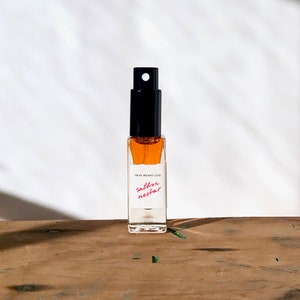 May include: A small glass bottle with a black spray top filled with a golden liquid. The bottle has a white label with the text "PETRA MORANE LOVE saffron nectar".