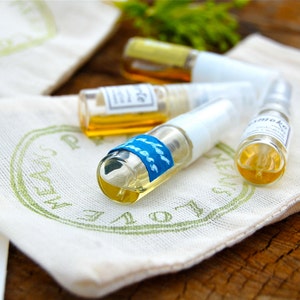 May include: Four small glass bottles with clear liquid and labels, sitting on a white fabric pouch with green lettering that says "LOVE ALWAYS".