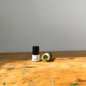 May include: Two small glass vials with black caps containing a pale yellow liquid. Each vial features a white label with the text "LITHA MEANS LOVE" and the handwritten script "Sainted rose" in red ink. The vials are positioned upright and on their side.