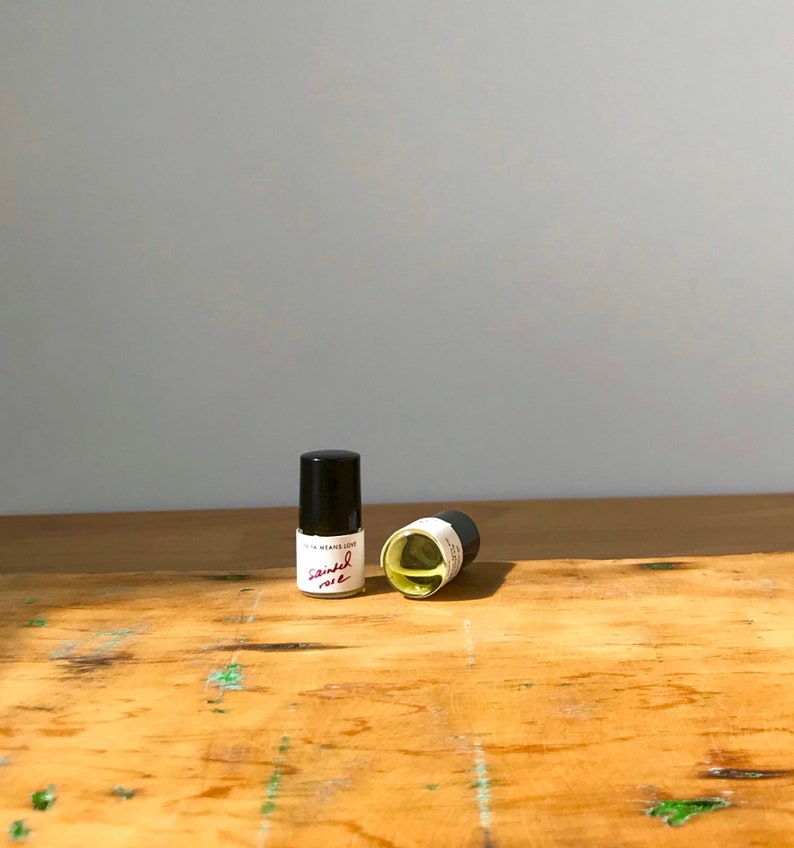 May include: Two small glass vials with black caps containing a pale yellow liquid. Each vial features a white label with the text "LITHA MEANS LOVE" and the handwritten script "Sainted rose" in red ink. The vials are positioned upright and on their side.