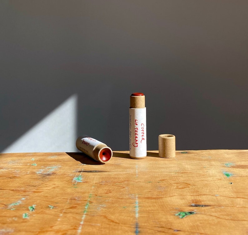 K&ouml;nnte beinhalten: Zwei braune Pappr&ouml;hrchen mit rotem Lippenbalsam. Das R&ouml;hrchen links ist mit "Pink" beschriftet und das R&ouml;hrchen rechts mit "Copper Lip Therapy".