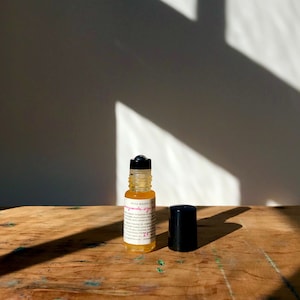 May include: A small glass bottle with a black rollerball top, filled with a yellow liquid. The bottle is labeled "Pomegranate eye serum" with a pink and white font. The bottle is sitting on a wooden surface.