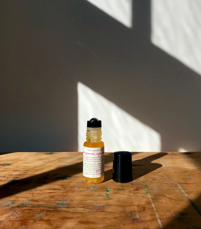 May include: A small glass bottle with a black rollerball top, filled with a yellow liquid. The bottle is labeled "Pomegranate eye serum" with a pink and white font. The bottle is sitting on a wooden surface.