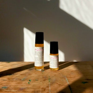 May include: Two glass roller bottles with black caps and labels. The labels are white with black text and pink writing that says "pomegranate oil". The bottles are sitting on a wooden surface.
