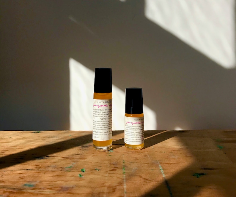 May include: Two glass roller bottles with black caps and labels. The labels are white with black text and pink writing that says "pomegranate oil". The bottles are sitting on a wooden surface.