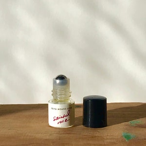 May include: A small clear glass roll-on perfume bottle with a silver metal ball applicator. The white label features the black text PRIYA MEANS LOVE and the red handwritten-style text Sainted rose. A separate black cylindrical cap sits beside it.