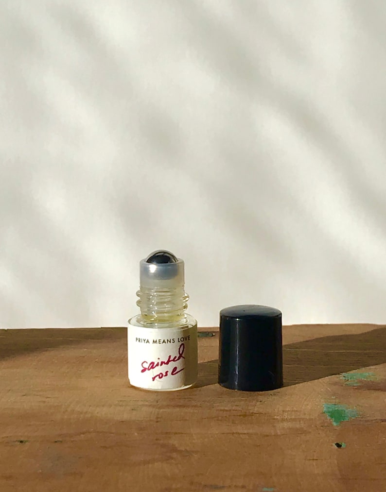 May include: A small clear glass roll-on perfume bottle with a silver metal ball applicator. The white label features the black text PRIYA MEANS LOVE and the red handwritten-style text Sainted rose. A separate black cylindrical cap sits beside it.