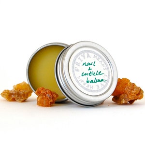 May include: A small silver tin with a white label that reads "Priya Means Love" and "nail & cuticle balm". The tin is open and a yellow balm is visible inside. There are three small pieces of amber-coloured resin on a white surface.