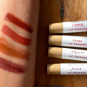 K&ouml;nnte beinhalten: Vier Tuben Lippenbalsam mit brauner Papierverpackung. Die Etiketten sind wei&szlig; mit schwarzem Text, der "PRIYA MEANS LOVE" und den Namen der einzelnen Geschmacksrichtungen lautet: "COPPER", "OCHRE", "CRIOLLO" und "MORA". Jedes Etikett tr&auml;gt au&szlig;erdem den Schriftzug "LIP THERAPY". Der Lippenbalsam ist in vier verschiedenen Braunt&ouml;nen auf einem Arm einer Person aufgetragen.