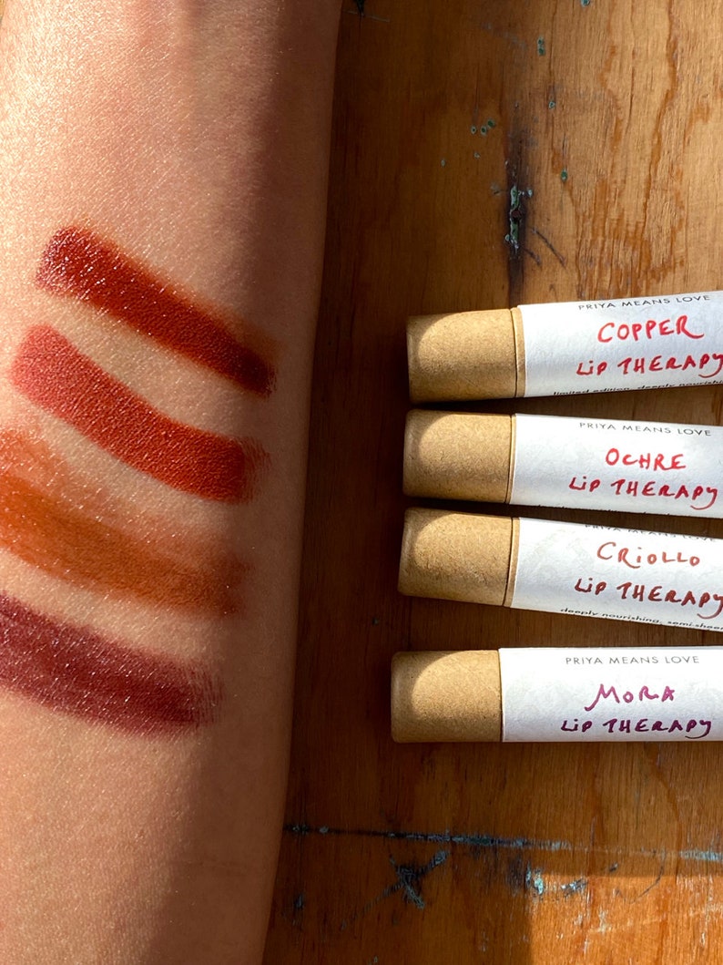K&ouml;nnte beinhalten: Vier Tuben Lippenbalsam mit brauner Papierverpackung. Die Etiketten sind wei&szlig; mit schwarzem Text, der "PRIYA MEANS LOVE" und den Namen der einzelnen Geschmacksrichtungen lautet: "COPPER", "OCHRE", "CRIOLLO" und "MORA". Jedes Etikett tr&auml;gt au&szlig;erdem den Schriftzug "LIP THERAPY". Der Lippenbalsam ist in vier verschiedenen Braunt&ouml;nen auf einem Arm einer Person aufgetragen.