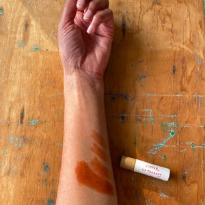 K&ouml;nnte beinhalten: Ein Arm mit einem Farbmuster in Braun, Orange und Rot. Eine kleine Pappr&ouml;hre mit dem Text "Copper Lip Therapy" liegt auf einer Holzoberfl&auml;che.