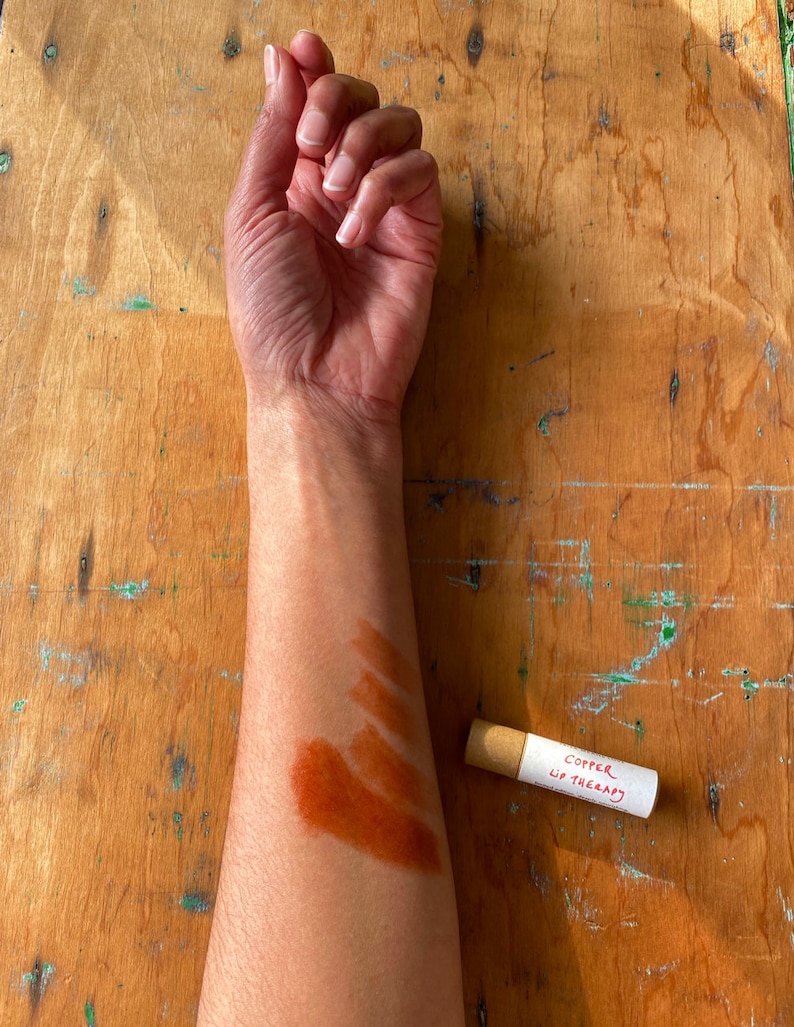 K&ouml;nnte beinhalten: Ein Arm mit einem Farbmuster in Braun, Orange und Rot. Eine kleine Pappr&ouml;hre mit dem Text "Copper Lip Therapy" liegt auf einer Holzoberfl&auml;che.