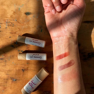 May include: Three tubes of lip balm with brown paper packaging. The tubes are labeled "Ochre Lip Therapy", "Mora Lip Therapy", and "Herbal Lip Therapy". The lip balm is shown swatched on a person's arm in three different shades: a light orange, a medium brown, and a light pink.