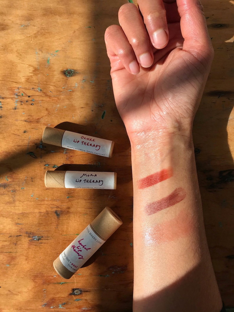 May include: Three tubes of lip balm with brown paper packaging. The tubes are labeled "Ochre Lip Therapy", "Mora Lip Therapy", and "Herbal Lip Therapy". The lip balm is shown swatched on a person's arm in three different shades: a light orange, a medium brown, and a light pink.