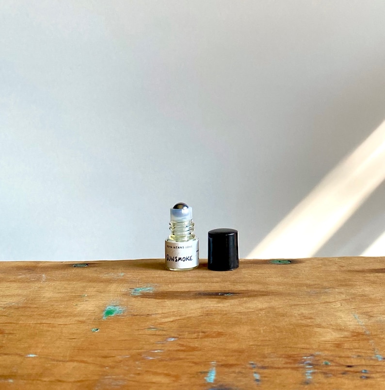 May include: A clear glass roller bottle with a silver roller ball and a black cap. The bottle is labelled "GUNSMOKE" with the text "WITH VITAMIN E OIL" underneath.