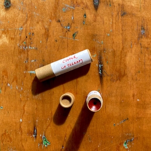 K&ouml;nnte beinhalten: Eine Pappr&ouml;hre mit einem wei&szlig;en Etikett, auf dem "Copper Lip Therapy" steht, und ein roter Lippenbalsam in einem Pappbeh&auml;lter.