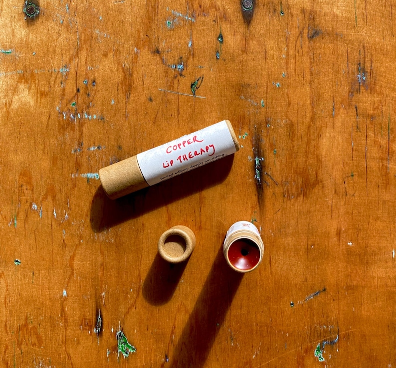 K&ouml;nnte beinhalten: Eine Pappr&ouml;hre mit einem wei&szlig;en Etikett, auf dem "Copper Lip Therapy" steht, und ein roter Lippenbalsam in einem Pappbeh&auml;lter.