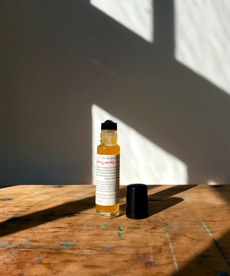 May include: A clear glass bottle with a black rollerball top filled with a yellow liquid. The bottle is labeled "PRIYA MEHRA" and "Pomegranate Serum". The bottle is sitting on a wooden surface.