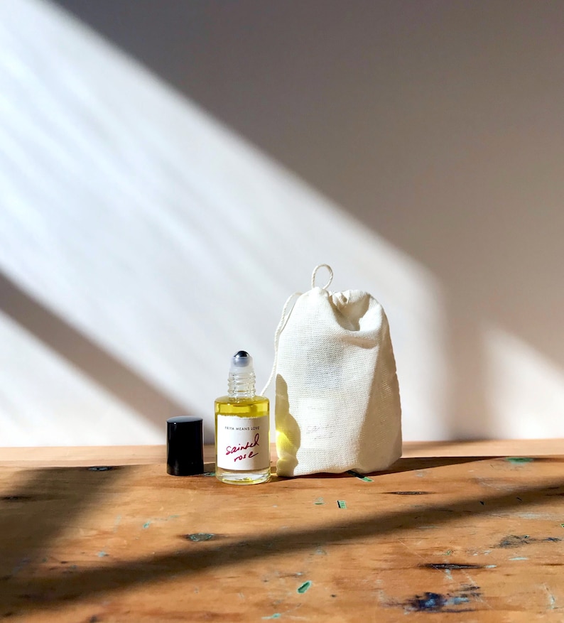 May include: A small clear glass rollerball bottle filled with yellow liquid, featuring a white label with the text "FRITA MEANS LOVE" and "sainted rose" in red script. A black cap sits beside the bottle, next to a small off-white drawstring pouch.