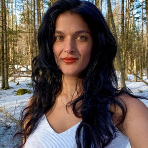 K&ouml;nnte beinhalten: Eine Frau mit langen, dunklen, welligen Haaren tr&auml;gt ein wei&szlig;es, gestreiftes Tanktop. Sie steht in einem schneebedeckten Wald mit hohen, kahlen B&auml;umen hinter ihr.