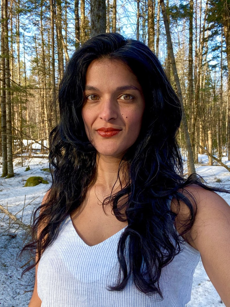 K&ouml;nnte beinhalten: Eine Frau mit langen, dunklen, welligen Haaren tr&auml;gt ein wei&szlig;es, gestreiftes Tanktop. Sie steht in einem schneebedeckten Wald mit hohen, kahlen B&auml;umen hinter ihr.