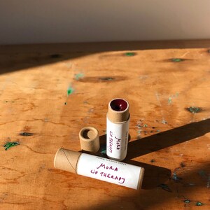 May include: Two brown cardboard tubes with white labels that say "Priya Means Love Mora Lip Therapy". The tubes are on a wooden surface. One tube has a red lip balm inside.