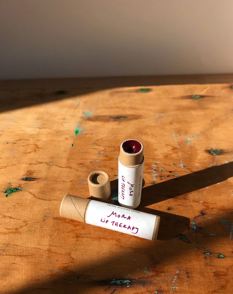 May include: Two brown cardboard tubes with white labels that say "Priya Means Love Mora Lip Therapy". The tubes are on a wooden surface. One tube has a red lip balm inside.