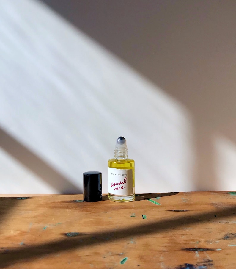 May include: A small clear glass roll-on bottle filled with a golden yellow liquid. The bottle features a white label with the text "FREYA MEANS LOVE" and "Sainted rose" written in red script. A black cylindrical cap sits next to the bottle.