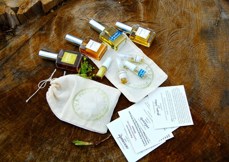 May include: A set of six small glass bottles of perfume, some with labels, on a wooden surface. The bottles are arranged in a circle, with a small white bag in the center. The bag has a green logo on it that says "Love & Made." There are also several small cards with text on them.