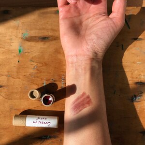 May include: A brown cardboard tube with the text "Mora Lip Therapy" on a white label. A brown lip balm is shown on a person's arm, with a brown smear of the balm on the skin.