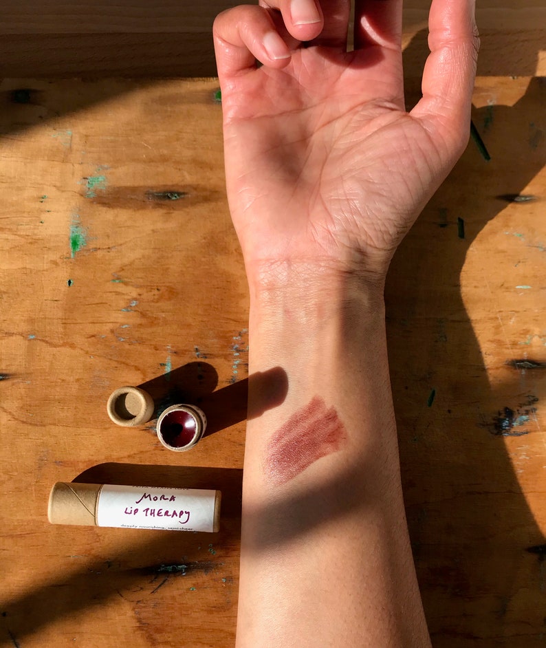 May include: A brown cardboard tube with the text "Mora Lip Therapy" on a white label. A brown lip balm is shown on a person's arm, with a brown smear of the balm on the skin.