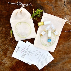 May include: Three small glass bottles of perfume in a white muslin bag with a green logo that says "Love, Me, Now". The bottles are labeled "Green Lilac", "Sun Smoke", and "Velvet Rose". There are also three small cards with descriptions of each scent.