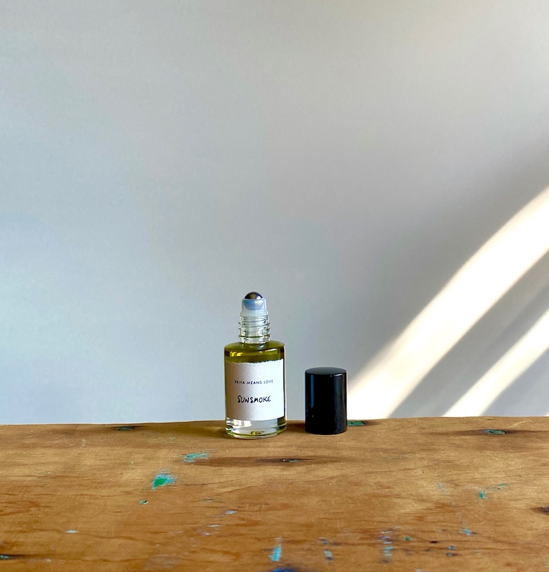 May include: A clear glass roller bottle with a black cap and a white label that reads "PETA MEANS LOVE SUNSMOKE". The bottle is sitting on a wooden surface.