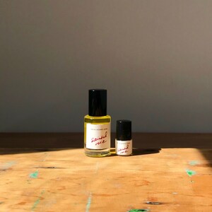 May include: Two clear glass bottles with black caps containing a yellow liquid. The labels are white with the text "FRITA MEANS LOVE" and "sainted rose" written in red script. The larger bottle is on the left and the smaller bottle is on the right.