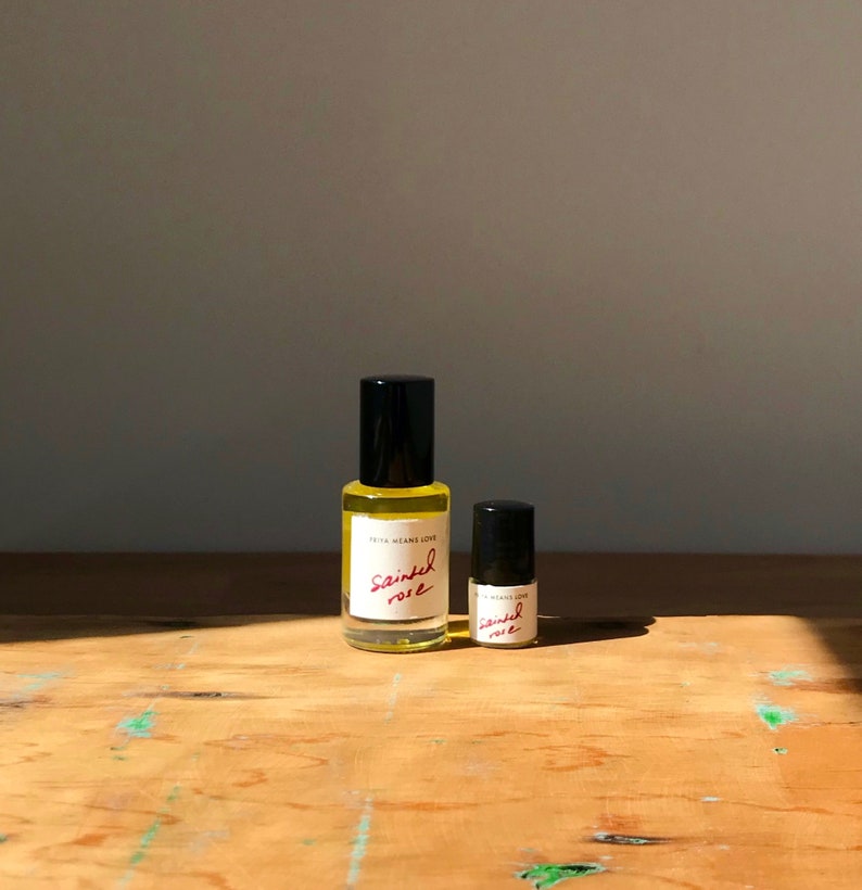 May include: Two clear glass bottles with black caps containing a yellow liquid. The labels are white with the text "FRITA MEANS LOVE" and "sainted rose" written in red script. The larger bottle is on the left and the smaller bottle is on the right.