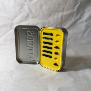 May include: A silver metal tin with the word "2010" embossed on the lid. Inside, a yellow insert holds several small, black rectangular objects. The tin is open, revealing the contents.