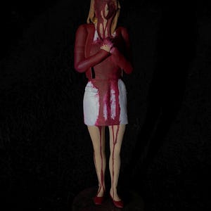 May include: A figurine of a nurse with blood-like paint covering the face, hair, and clothing. The nurse is wearing a white cap, red jacket, white skirt, and red shoes. The blood-like paint drips down the figurine.
