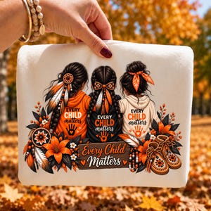 May include: A white sweatshirt featuring a graphic of three individuals with braided hair, wearing orange and white hoodies. The design includes the text Every Child Matters, orange flowers, feathers, a dreamcatcher, and beaded moccasins.
