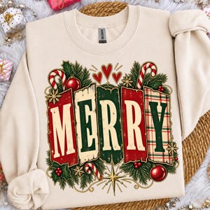 Könnte beinhalten: Ein cremefarbenes Sweatshirt mit Rundhalsausschnitt und einem Grafikdruck des Wortes MERRY in großen Blockbuchstaben. Das Design umfasst rote und grüne Felder, Zuckerstangen, Stechpalmenblätter, rote Ornamente, goldene Sterne und kleine rote Herzen.